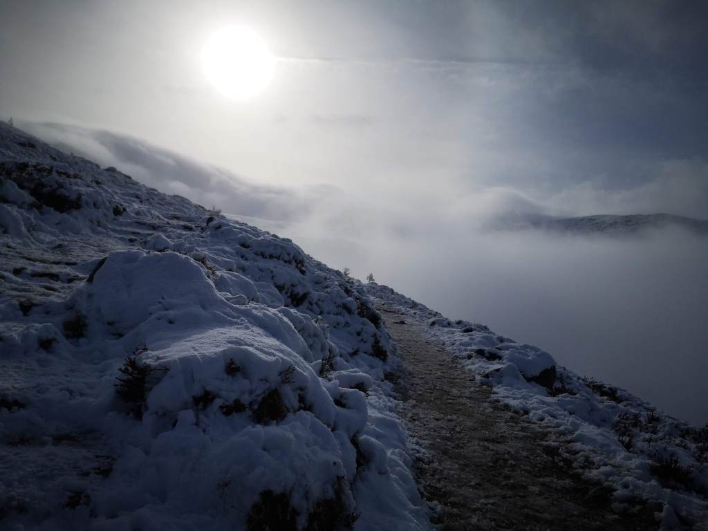 Image of a snow covered mountain side I stood on the path while taking it so the path emerges from the foreground disappearing almost immediately behind a bend. The horizon is thick snowclouds but just above is a bit of blue sky and the winter sun has a huge halo around it. 

CC Nathalie Tasler 